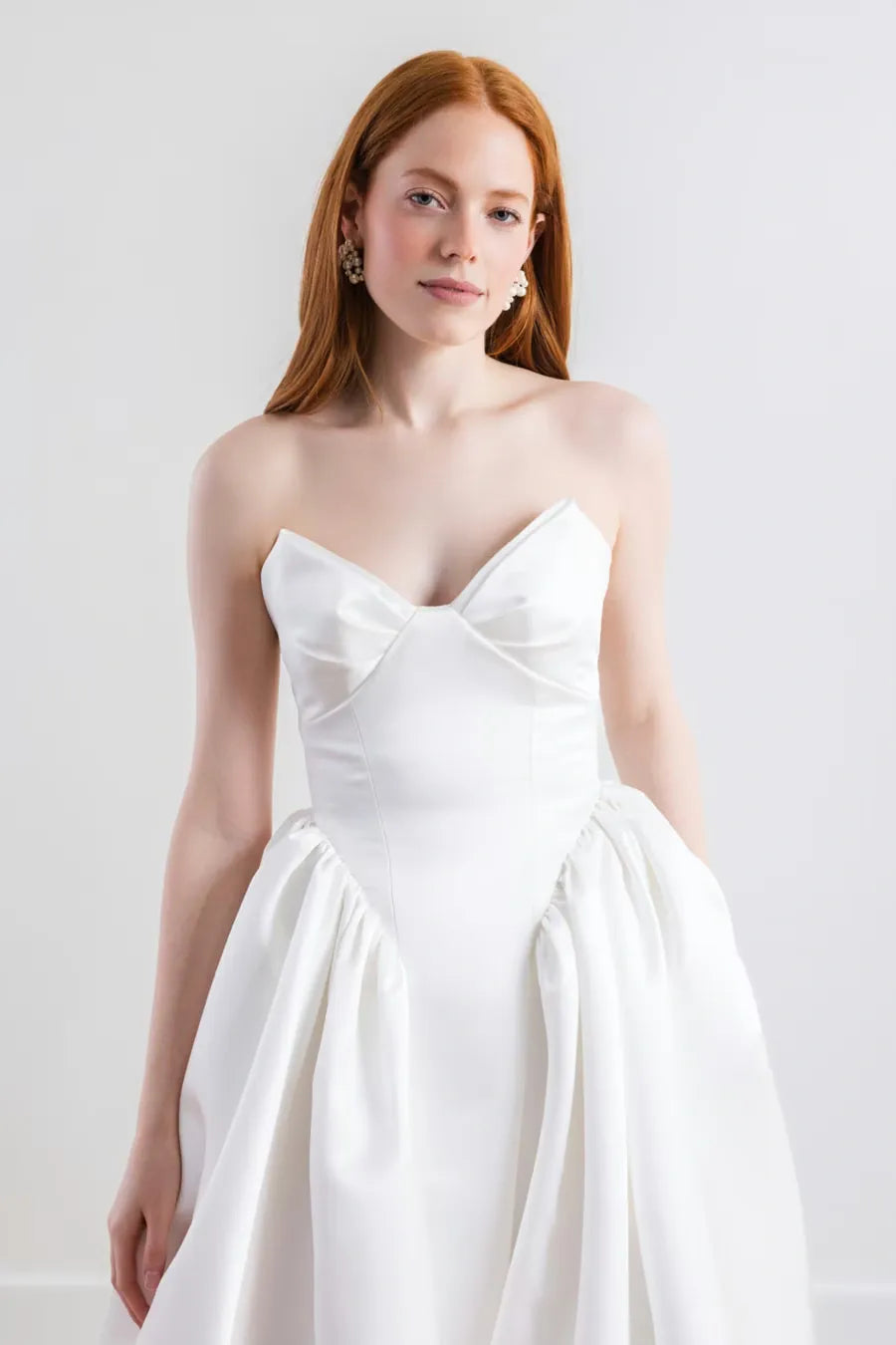 A red-haired woman wearing a strapless white satin gown with a sweetheart neckline, bow detail at the bust, and a voluminous skirt, photographed against a plain light gray studio background.