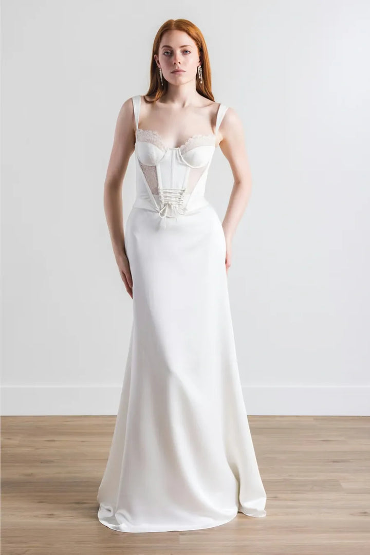 A woman modeling a floor-length white wedding gown with a structured corset-style bodice, sheer mesh panels, and a smooth satin skirt, standing against a plain light wall on a wood floor.
