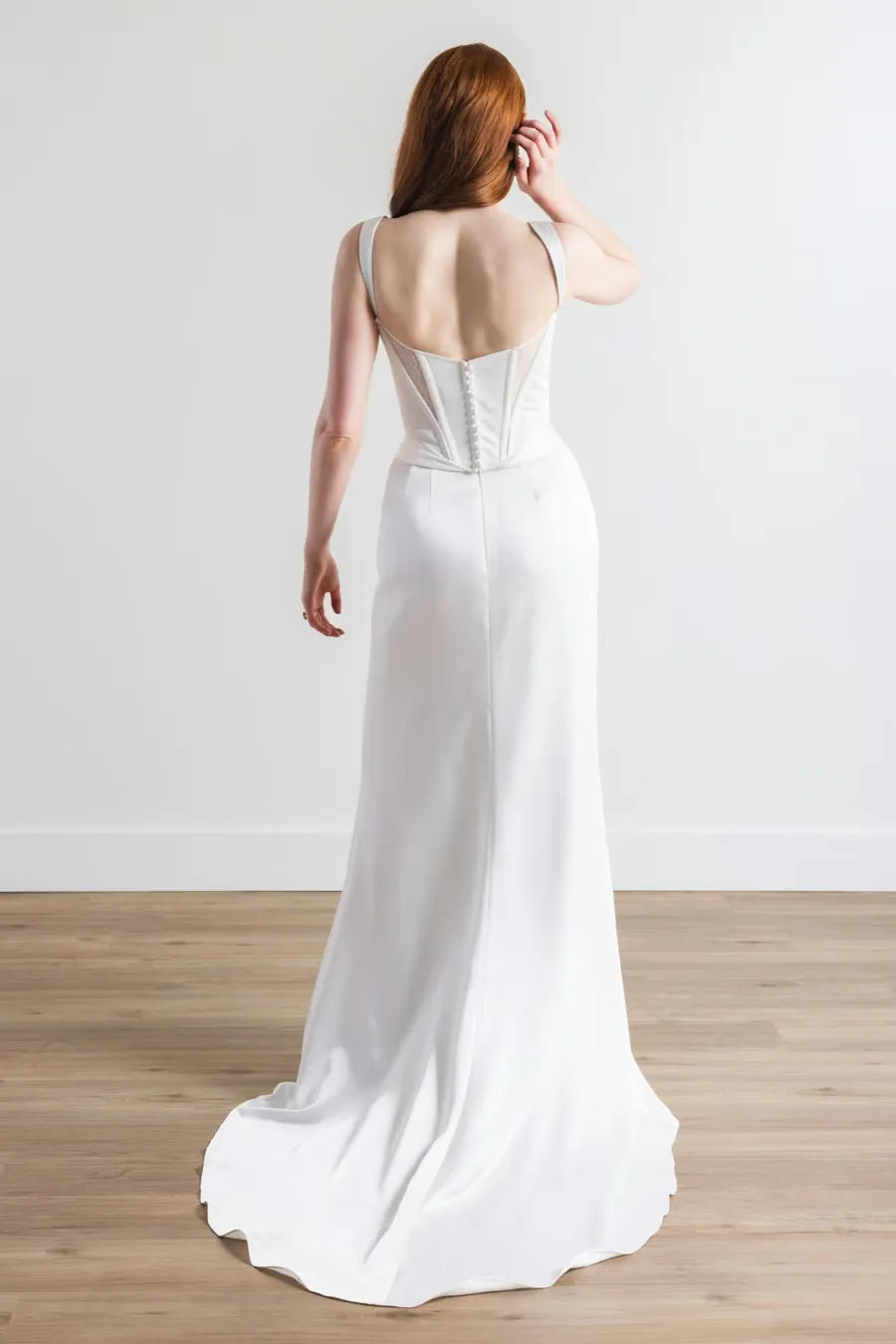A woman wearing a white sleeveless wedding gown is shown from the back in a bright indoor room with a light wall and wood floor. The dress has a fitted corset-style back with lace-up detailing and a flowing train.