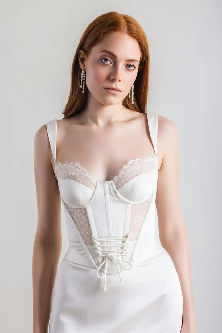 A woman with red hair wearing a white corset-style dress with lace trim and a lace-up front, photographed against a plain light gray studio background.