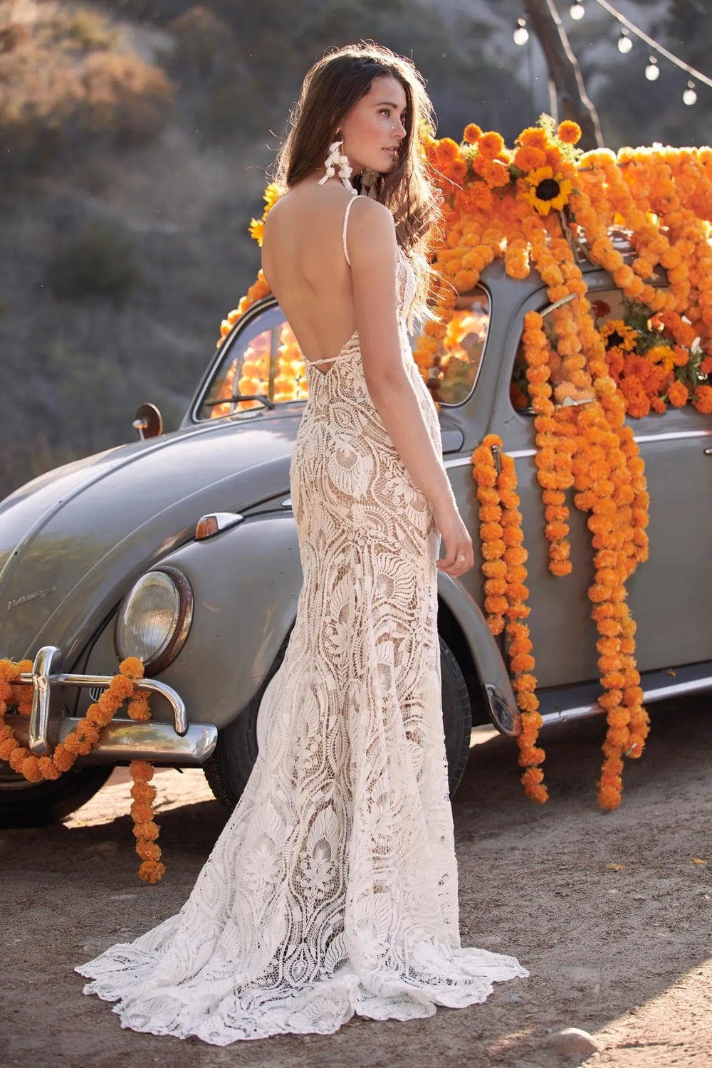 Woman in a white lace dress standing next to a vintage car decorated with orange flowers.