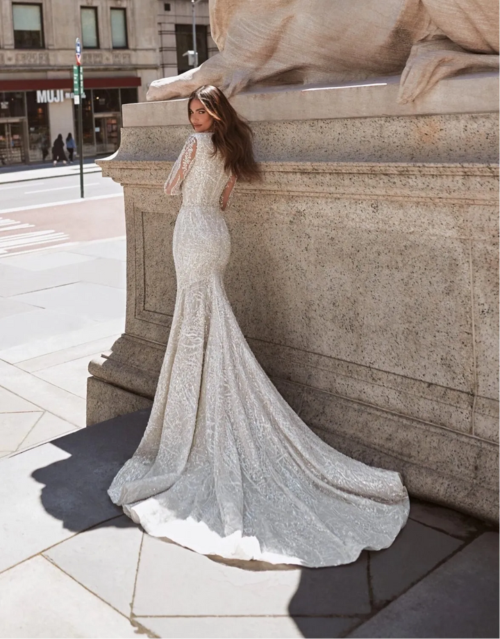 A woman wearing a sparkling silver mermaid-style wedding gown with a long train stands beside a large stone lion statue in an urban city setting.
