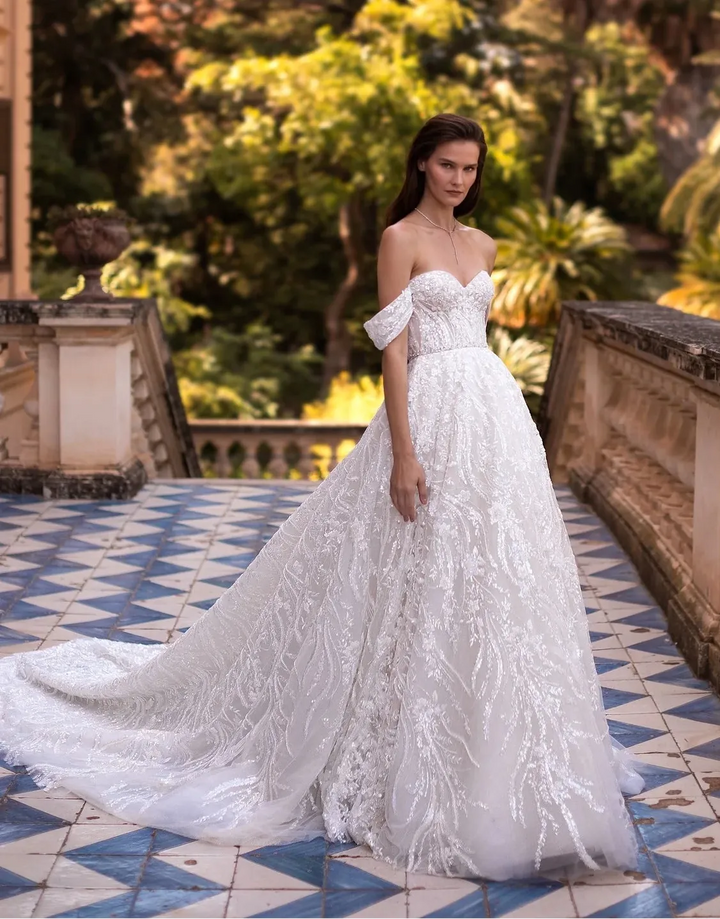 A woman wearing a white off-the-shoulder lace wedding gown stands on a tiled terrace with a lush garden background.