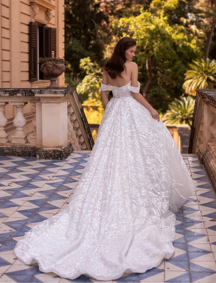 A woman wearing a voluminous white lace wedding gown stands on a tiled terrace with a historic building and lush green trees in the background.