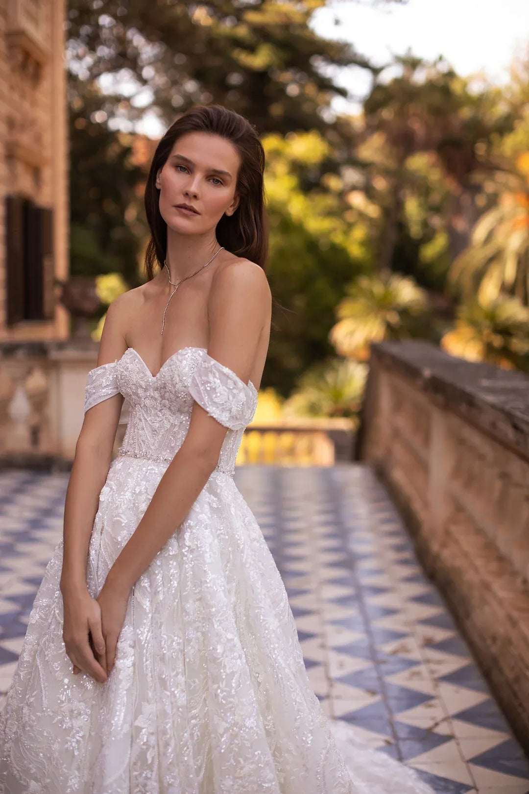 A woman wearing an off-the-shoulder white lace wedding gown stands on a tiled outdoor walkway with a blurred stone building and greenery in the background.