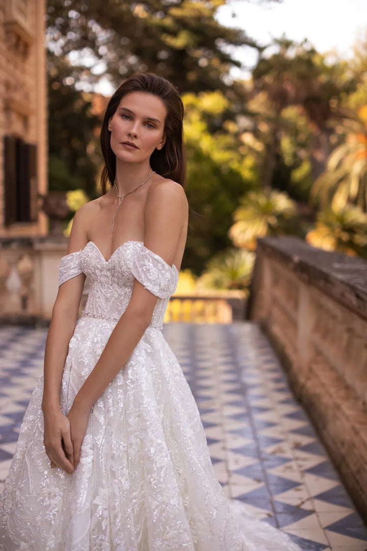 A woman wearing an off-the-shoulder white lace wedding gown stands on a tiled outdoor walkway with a blurred stone building and greenery in the background.