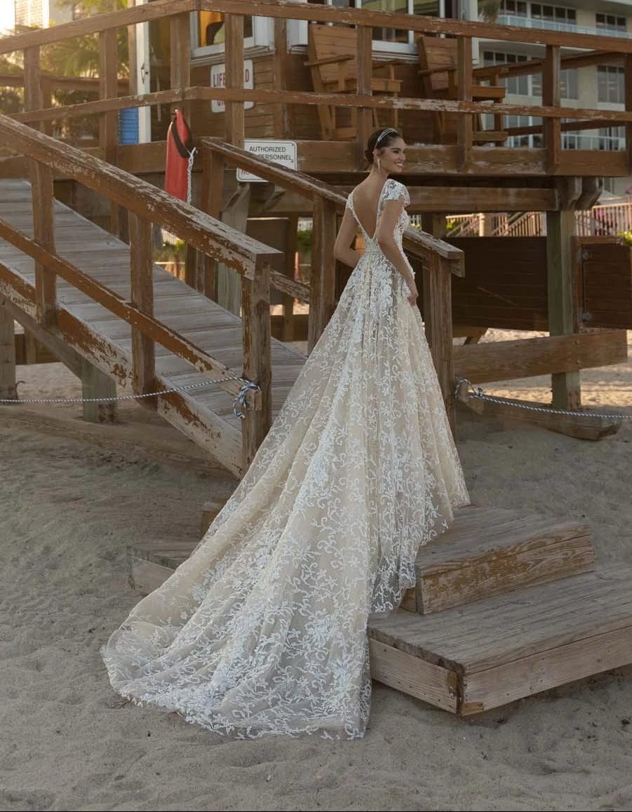 Woman in a wedding dress standing on a sandy beach near a wooden gazebo with buildings in the background.