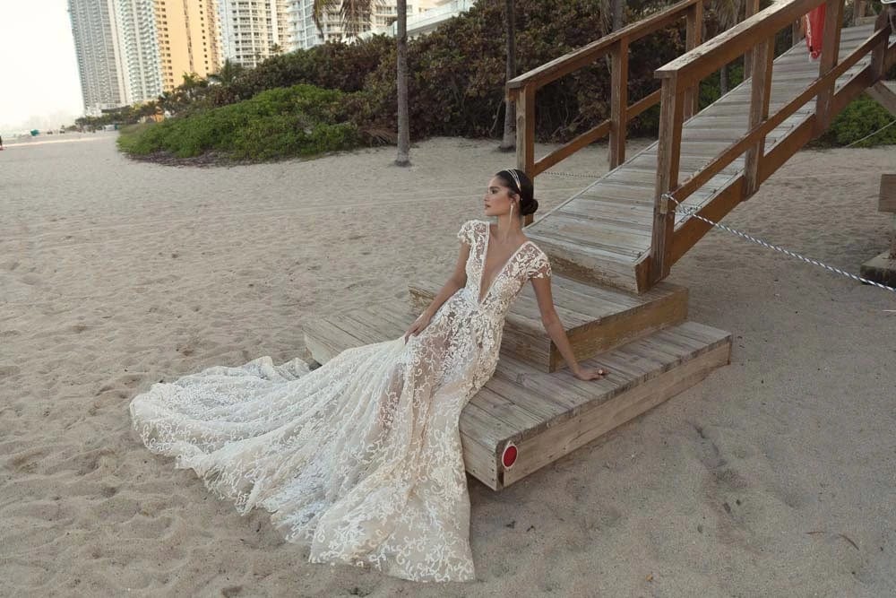 Woman in a white wedding dress sitting on a wooden bench by the beach.