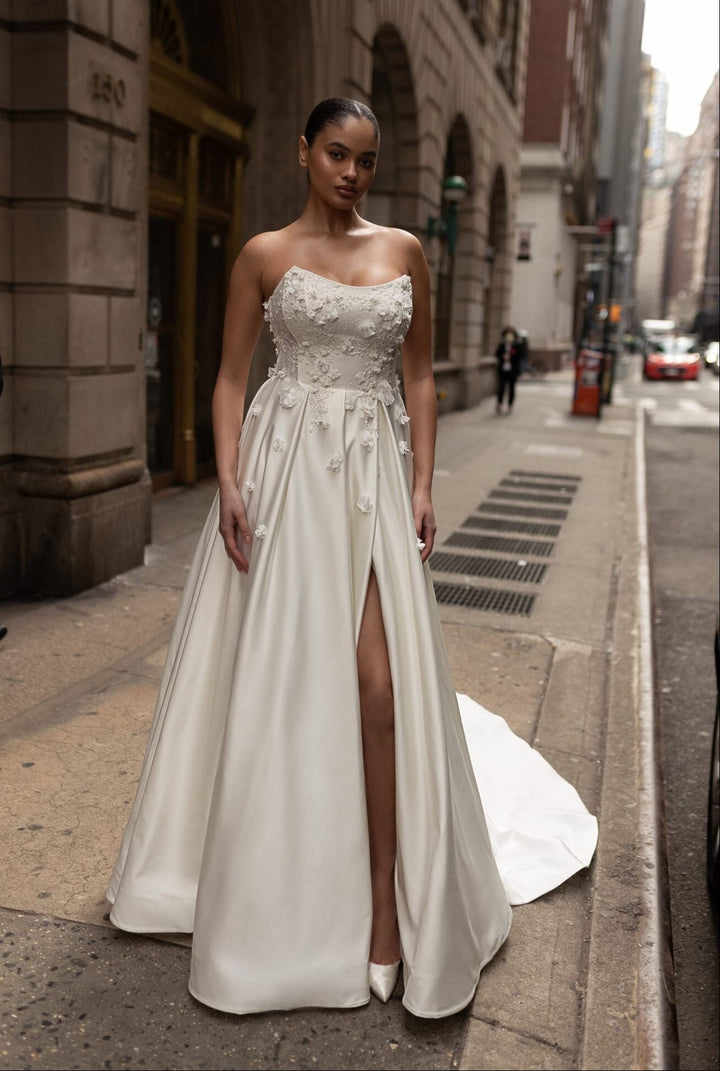 A woman wearing a strapless ivory wedding gown with a fitted bodice, sheer lace overlay, and a high front slit, standing on a city sidewalk.