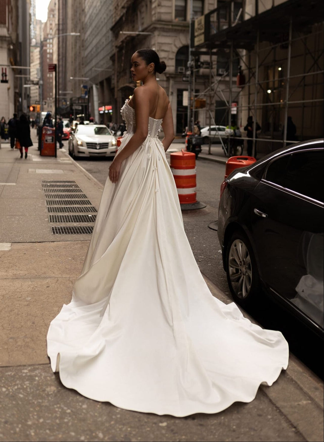 A woman wearing a strapless white wedding gown with a full skirt and long train stands on a city street, photographed from behind as she looks back over her shoulder. The background shows urban buildings, parked cars, and pedestrians.