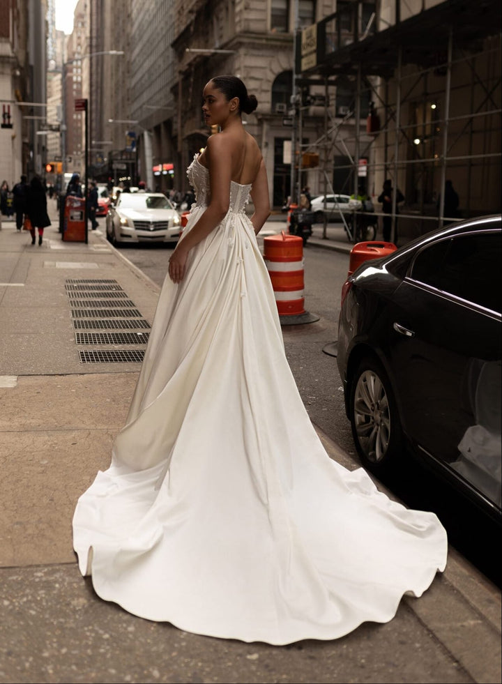 A woman wearing a strapless white wedding gown with a full skirt and long train stands on a city street, photographed from behind as she looks back over her shoulder. The background shows urban buildings, parked cars, and pedestrians.