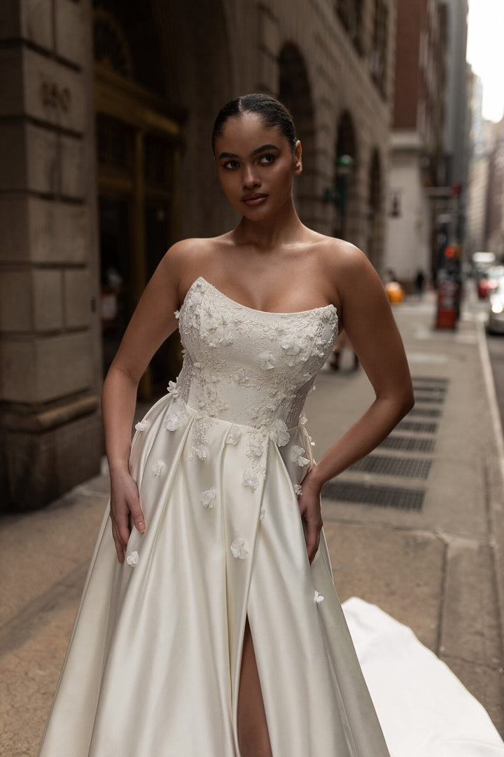 A woman modeling a strapless ivory wedding gown with 3D floral applique on the bodice and waist, standing on a city street.