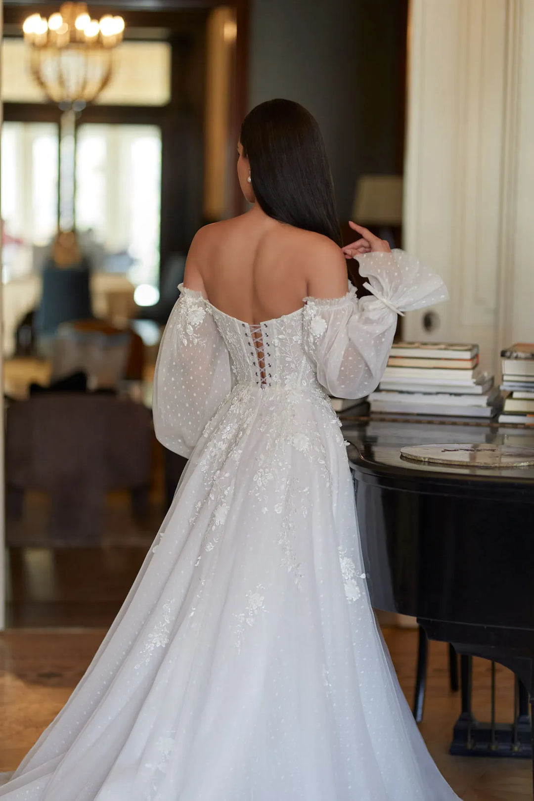 Woman in a white lace dress standing in an elegant room with a chandelier.