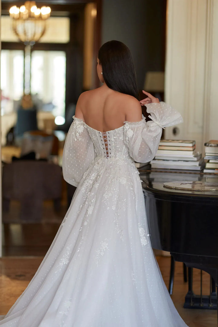 Woman in a white lace dress standing in an elegant room with a chandelier.
