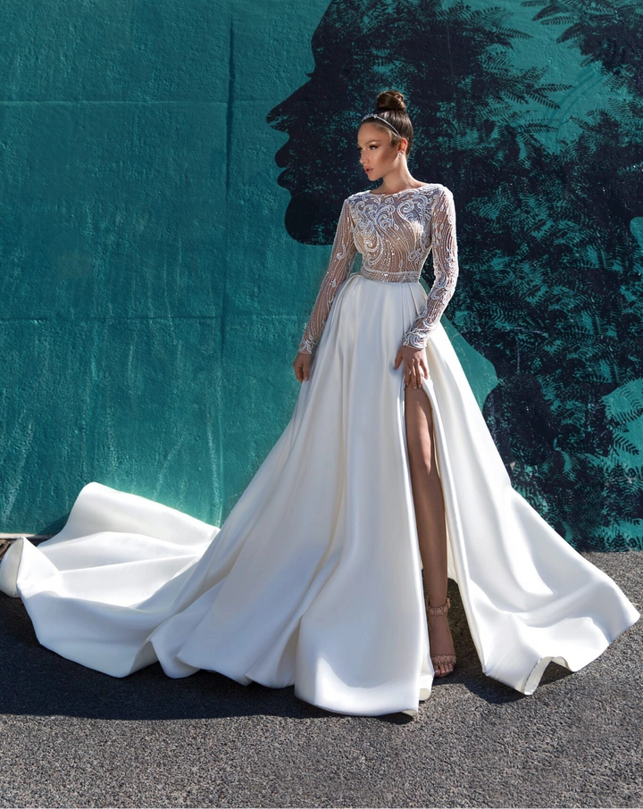 A woman models a floor-length white evening gown with a high thigh slit, sheer lace long sleeves, and a structured bodice, standing in front of a dark teal backdrop with large white lettering.