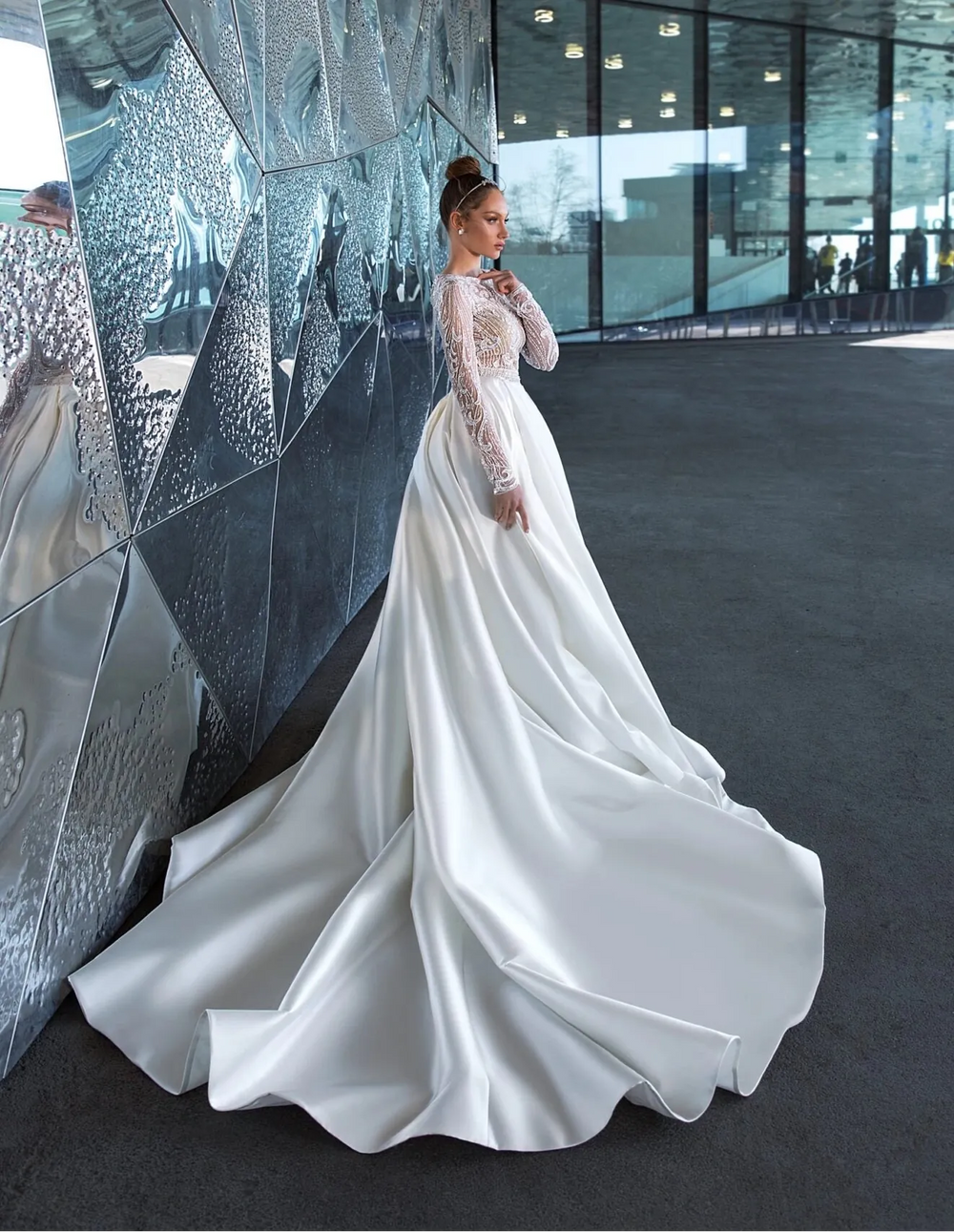 A woman wearing a long-sleeved white satin wedding gown with lace sleeves, standing in a modern interior space with reflective metallic walls.