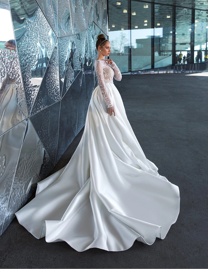 A woman wearing a long-sleeved white satin wedding gown with lace sleeves, standing in a modern interior space with reflective metallic walls.