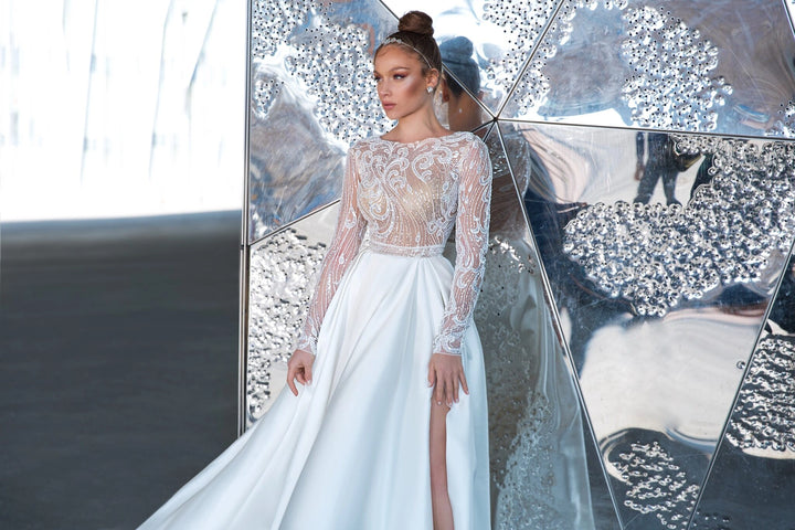 A woman models a white wedding gown with a sheer lace bodice, long sleeves, and a high slit, standing in front of reflective mirrored panels.