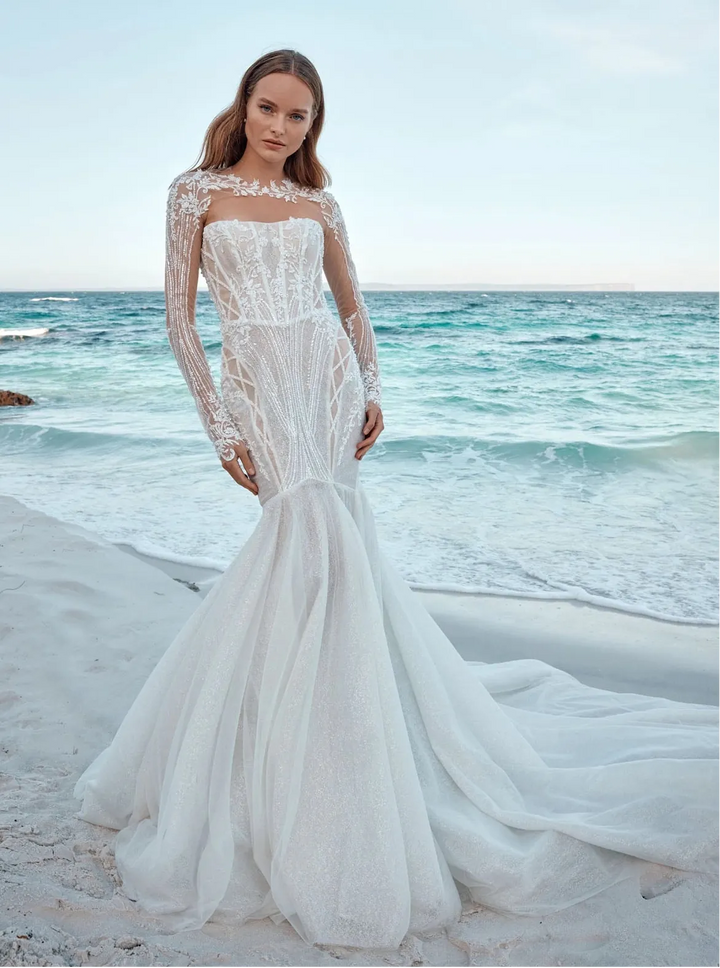 Woman in a white lace wedding dress standing on a beach with ocean in the background