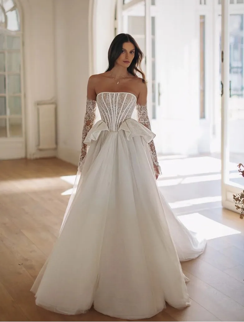 Woman in a white strapless gown with long sleeves standing in a bright room with large windows.