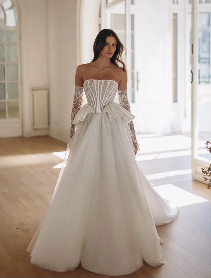 Woman in a white strapless gown with long sleeves standing in a bright room with large windows.
