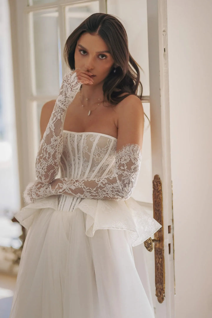 Woman wearing a white lace dress with sheer sleeves in a softly lit room.