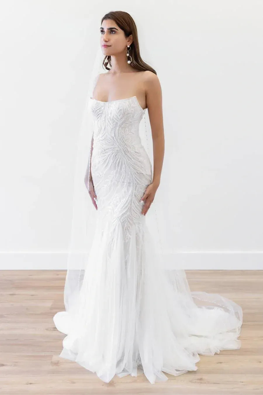 A woman modeling a strapless white mermaid-style wedding gown with a fitted lace bodice, sheer tulle skirt, and long flowing train against a plain white wall and light wood floor.
