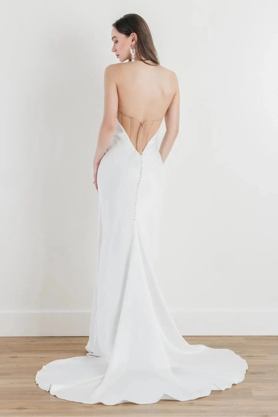 A woman modeling a white bridal gown from the back, showing a deep open back and train, standing on a light wood floor against a white wall.