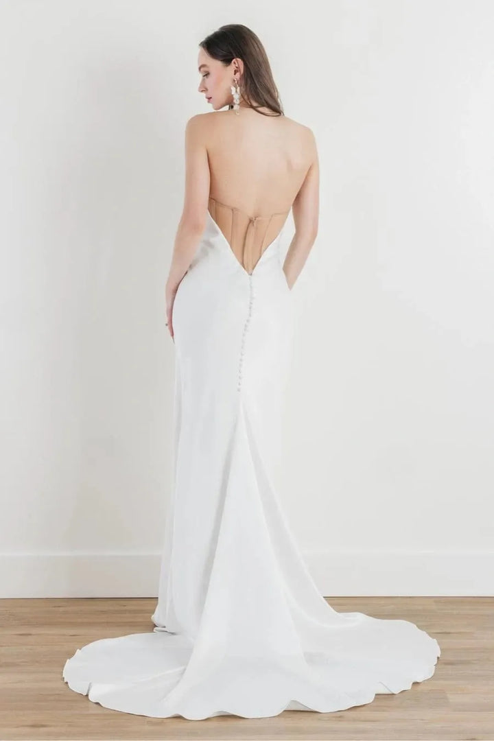 A woman modeling a white bridal gown from the back, showing a deep open back and train, standing on a light wood floor against a white wall.
