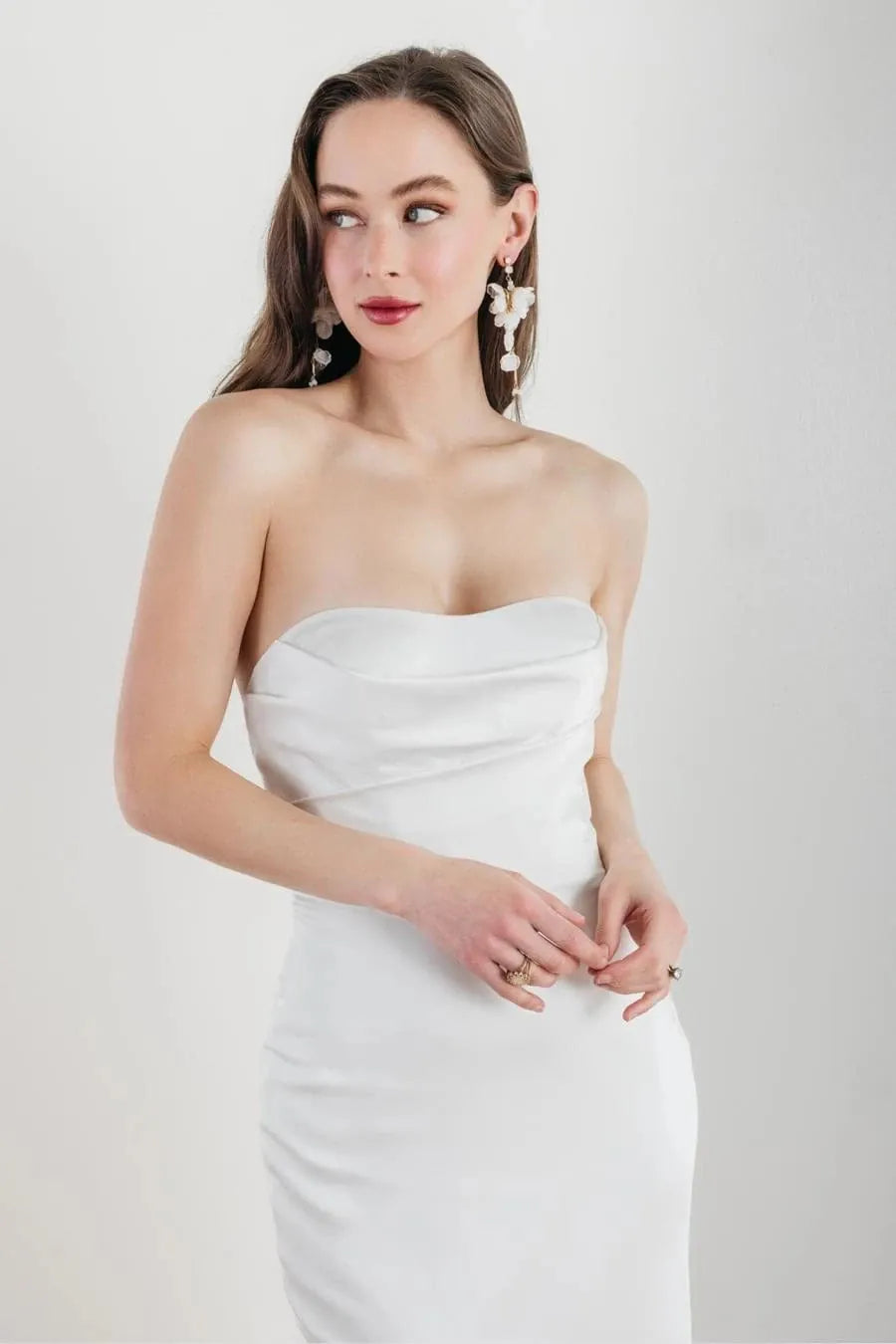 A woman wearing a strapless white fitted dress with a draped sweetheart neckline, photographed against a clean light gray studio background.