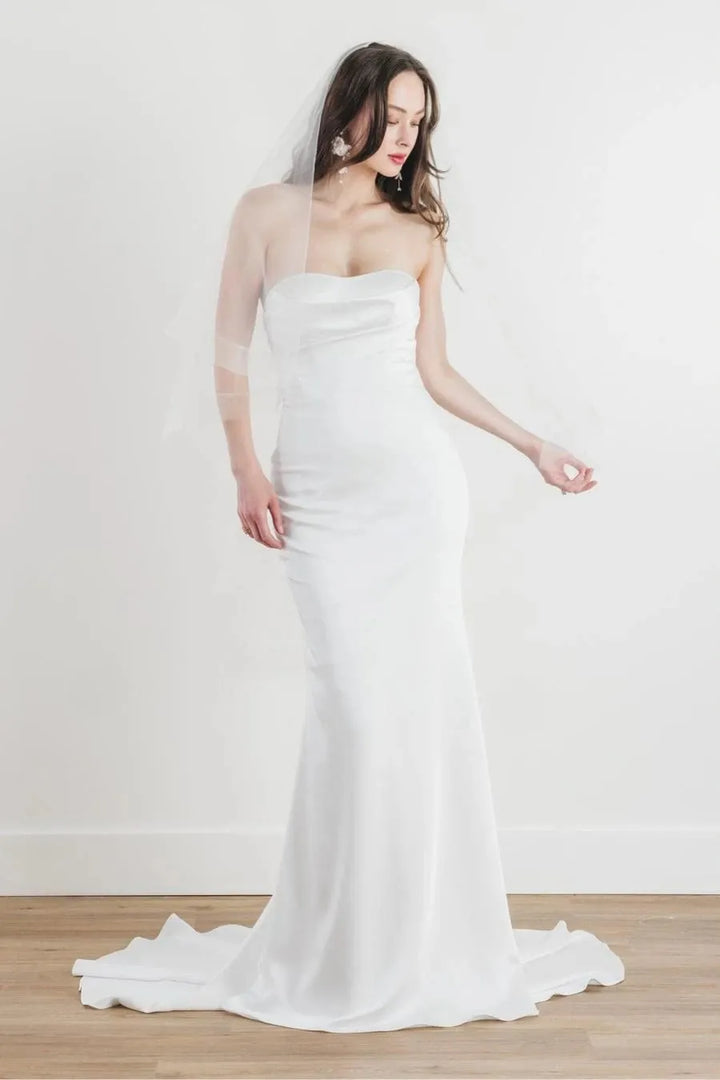 A woman modeling a strapless white wedding gown with a fitted silhouette, sheer long sleeves, and a long train, standing against a plain light wall on a wood floor.