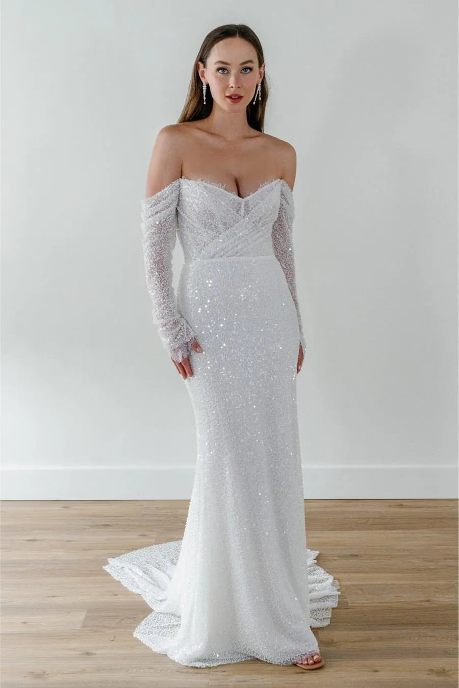 A woman modeling an off-the-shoulder white wedding gown covered in sequins, featuring long sheer sleeves and a fitted mermaid silhouette with a train, standing on a light wood floor against a white wall.